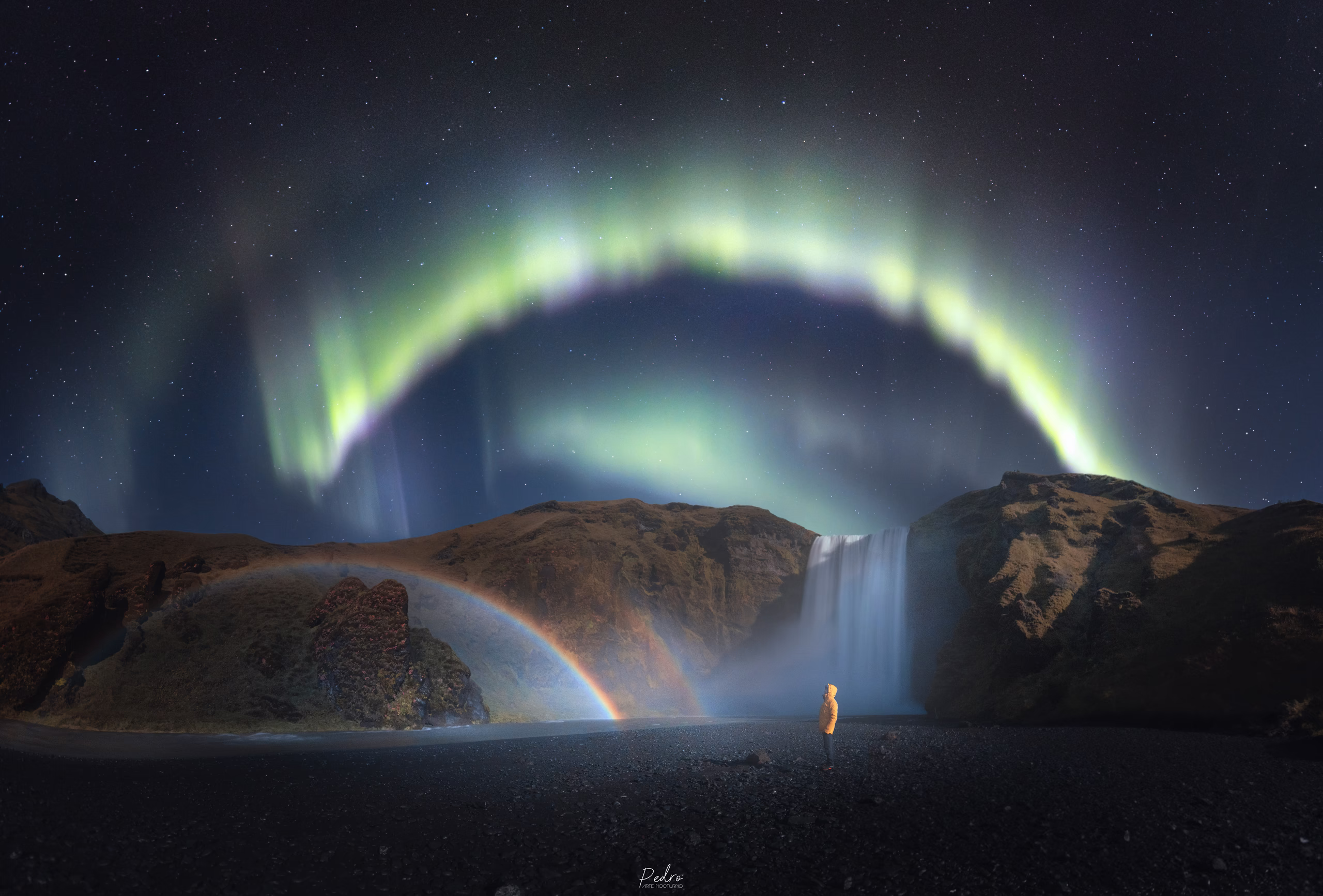 Una cascada bajo una aurora boreal, con un arcoíris en el cielo nocturno.